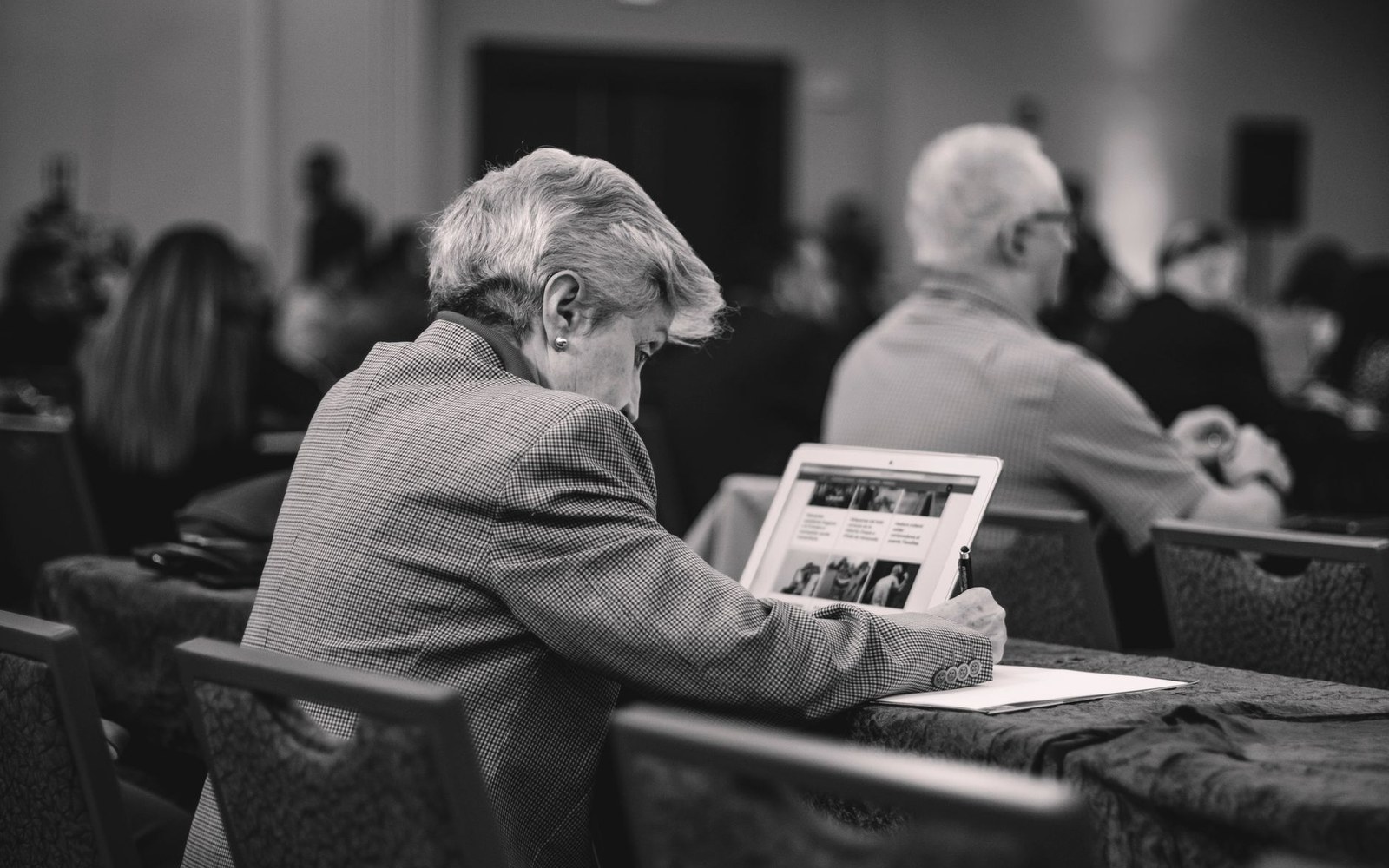 A seated attendee looks down at a tablet while others sit in rows nearby