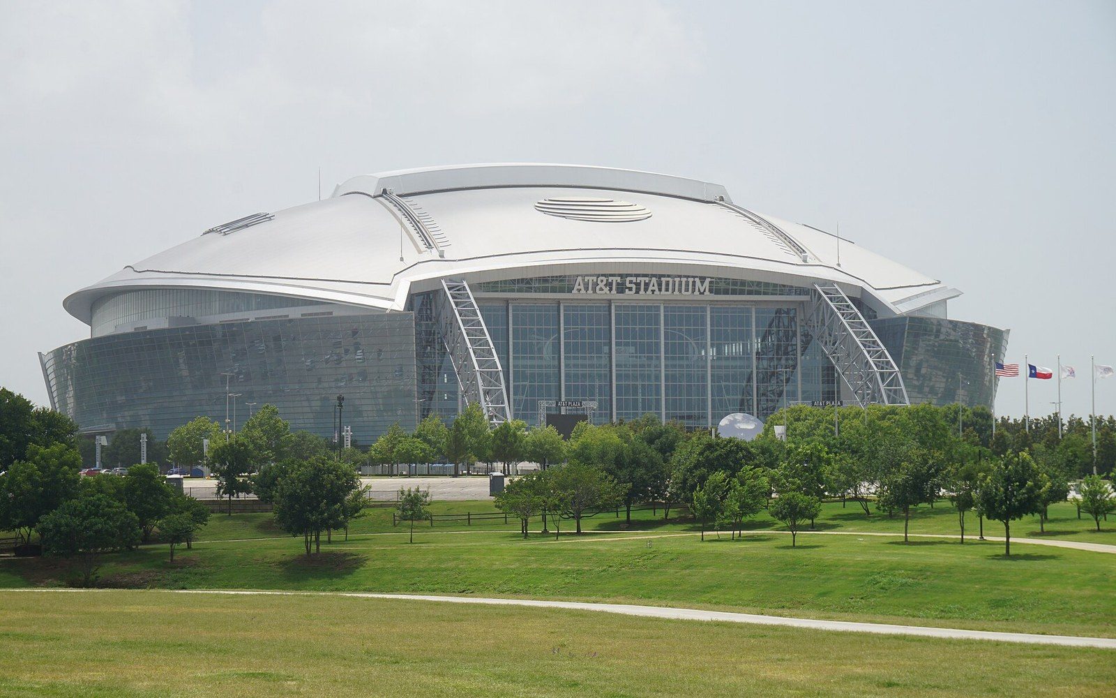 AT&T Stadium's curved glass facade rises behind a grassy park with scattered trees