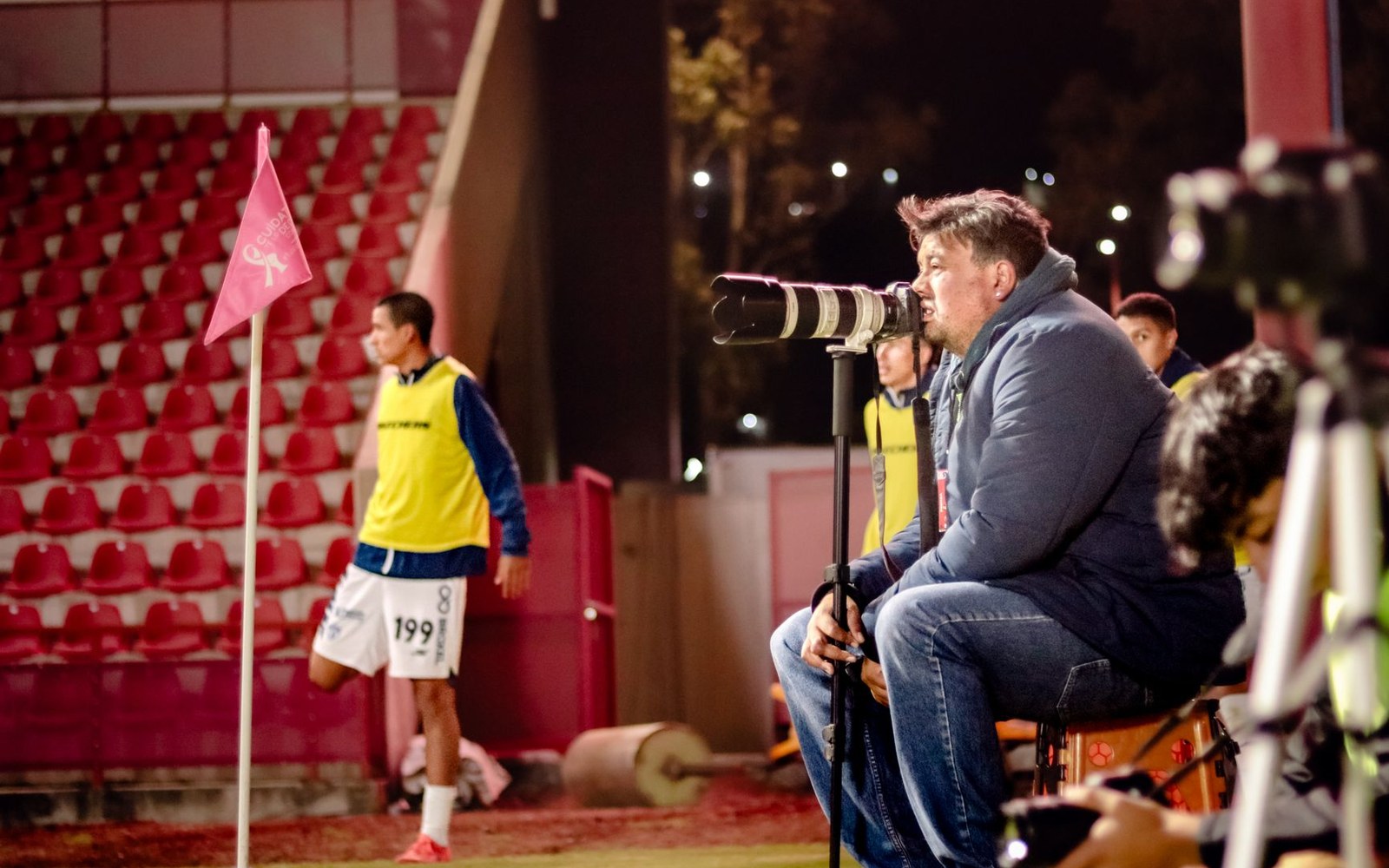A photographer with a large telephoto lens sits pitchside as a footballer warms up nearby
