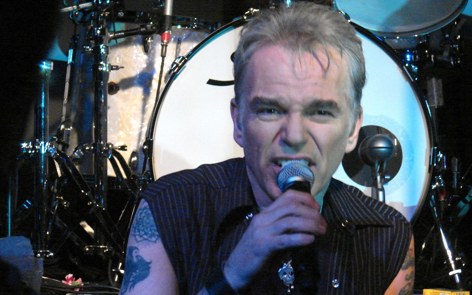 Tattooed singer with grey hair grips a microphone in front of a drum kit